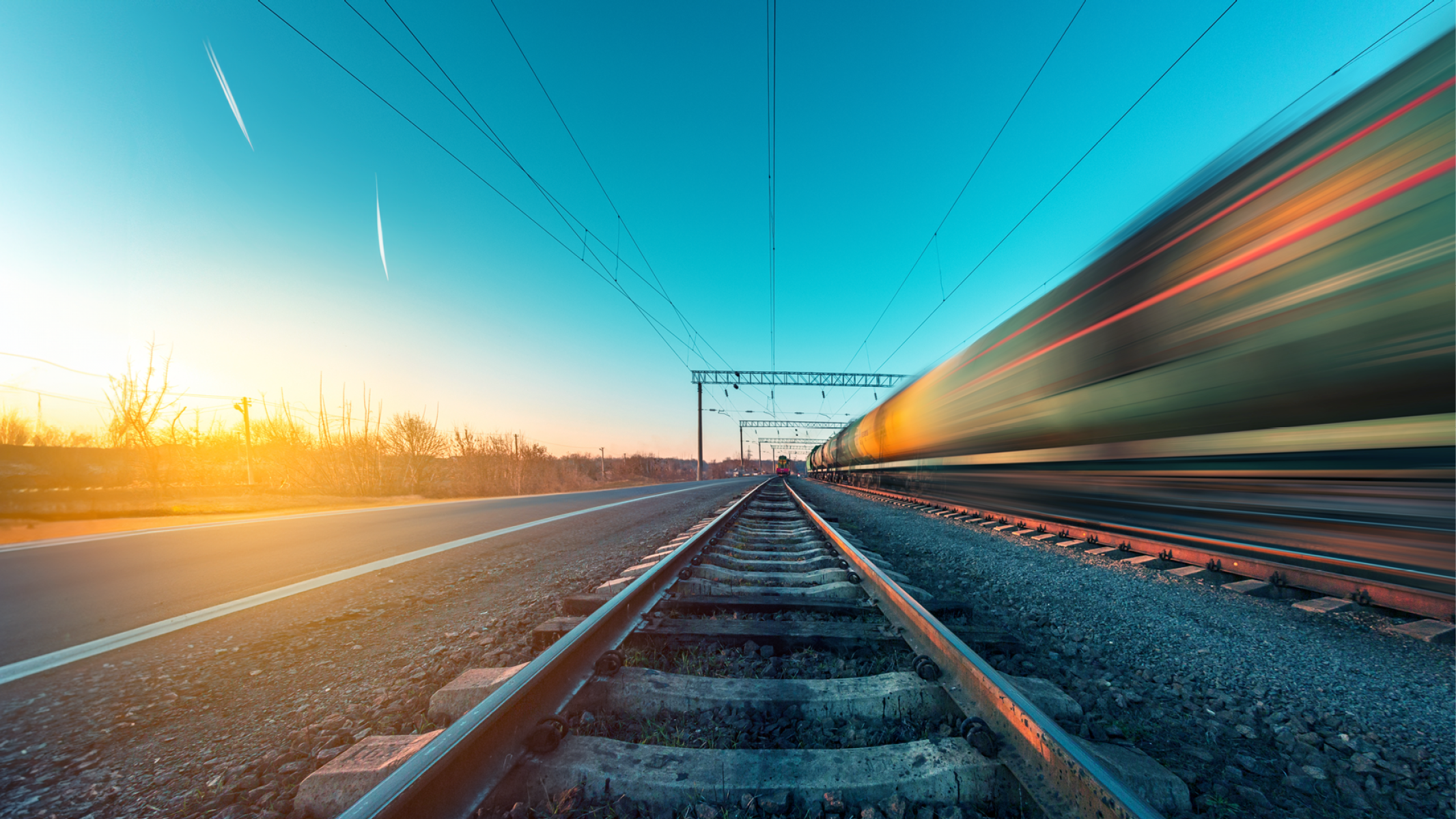 Binario ferroviario al tramonto con un treno in movimento sfocato che passa accanto a una strada asfaltata. Descrizione generata da AI